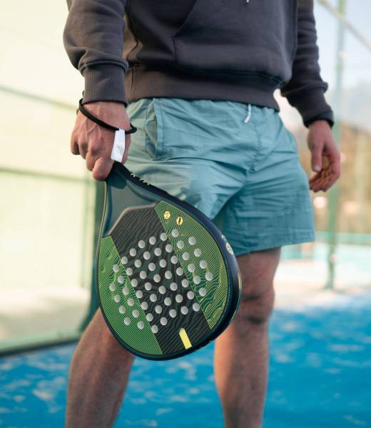 Close-up of a man's hands gripping a padel tennis racket, ready to play, with the court's fence and blue surface in the background. High quality photo