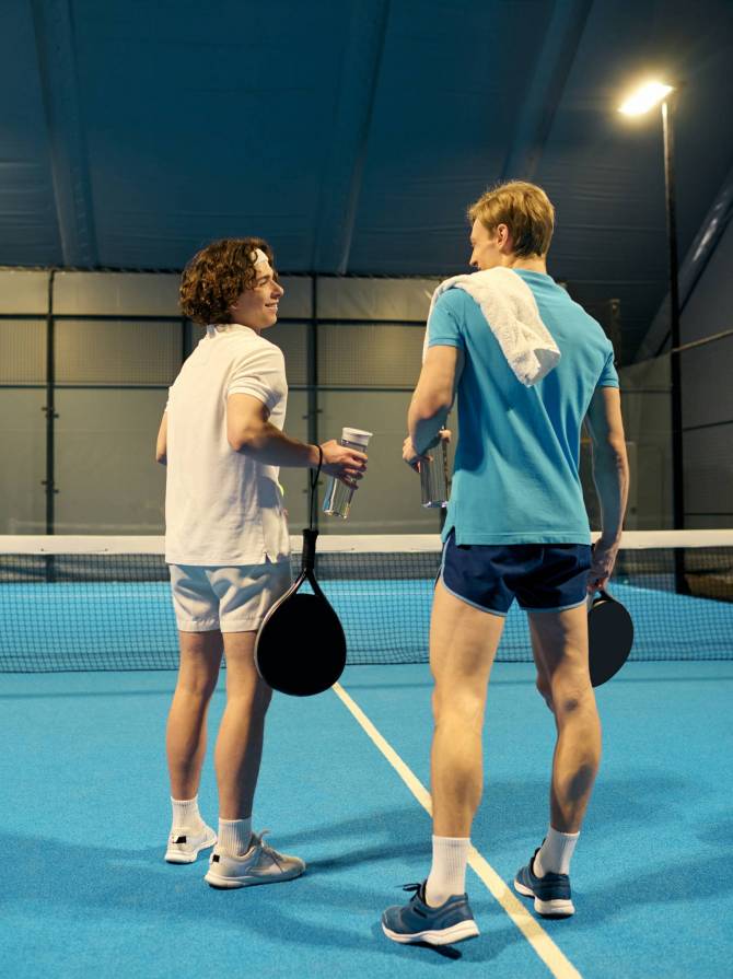 Two young sportsmen pause to chat and hydrate after an energetic padel match on a bright court.